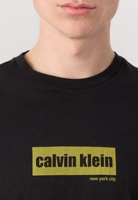Svart bomull T-skjorte med en gul rektangulær logo som har teksten "calvin klein" og "new york city" i fet, fremtredende skrift.