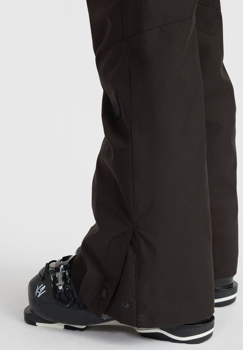 Braune Schneehosen aus leicht strukturiertem Stoff, mit seitlichen Reißverschlüssen und einer weiteren Passform an den Knöcheln, kombiniert mit schwarzen Skischuhen.