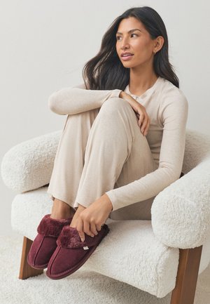 Vrouw zittend in een witte, gestructureerde fauteuil, gekleed in beige loungewear en kastanjebruine bontpantoffels, kijkt bedachtzaam opzij.