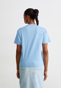 Maglietta in cotone azzurro chiaro con maniche corte e collo rotondo, vista da dietro. Indossata con una gonna in denim chiaro e grandi orecchini a cerchio in argento.