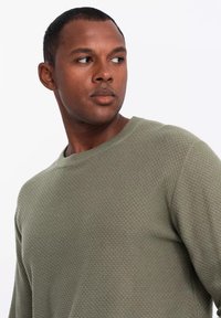 Maglione verde con texture, collo a giro, polsini a coste e una vestibilità rilassata, indossato da una persona che guarda di lato.