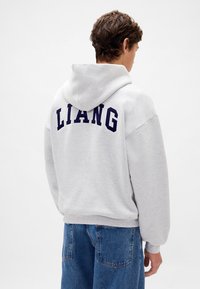 Szara bluza z kapturem z dużym granatowym napisem „LIANG” na plecach. Wykonana z bawełny, posiada kaptur ze sznurkiem i prążkowane mankiety.