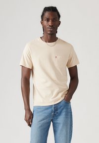 Beige bomulds T-shirt med rund hals og lomme på venstre bryst, parret med lyseblå denim jeans. Modellen har tekstureret hår og en kædehalskæde.