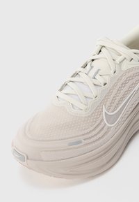Beżowe sneakersy Nike z siateczkową teksturą, białymi sznurówkami i widocznym logo Nike na boku i palcu, sfotografowane na białym tle.