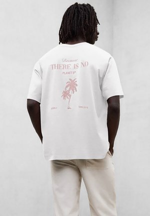 Man met dreadlocks die van achteren te zien is, draagt een wit T-shirt met roze palmboompjes en de tekst "ER IS GEEN PLANEET B" op de achterkant, beige broek.
