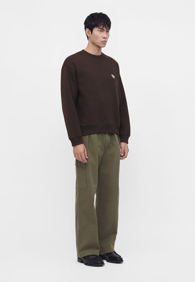 Jeune homme debout portant un sweat-shirt marron foncé avec un petit logo et un pantalon cargo ample olive avec des chaussures noires.