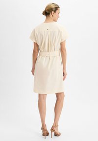 Frau mit blondem, zu einem Dutt gebundenem Haar, trägt ein cremefarbenes, kurzärmeliges Kleid mit Gürtel und nude-farbene High Heel Sandalen, steht mit dem Rücken zur Kamera.