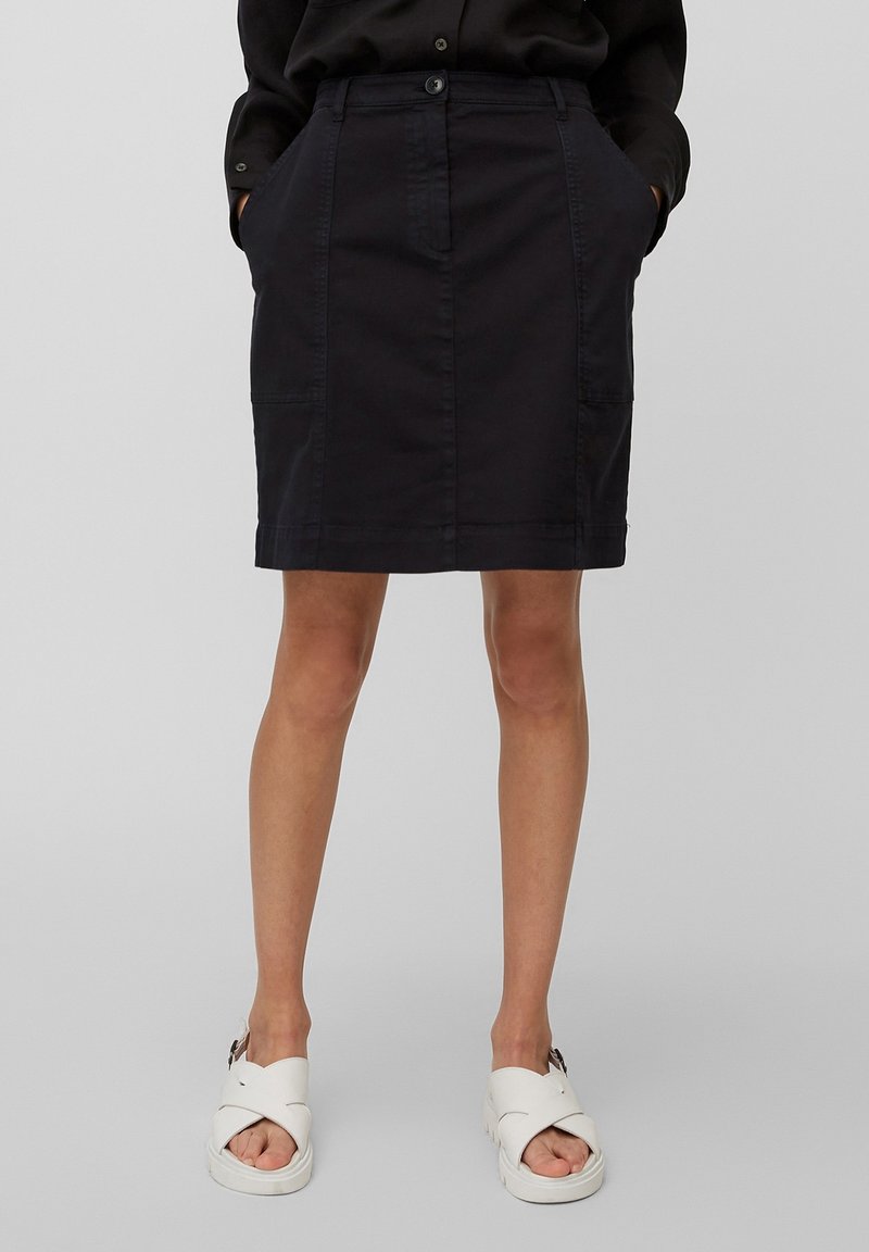 Femme portant une jupe noire mi-longue avec des poches à l'avant, associée à des sandales blanches à bouts ouverts, debout devant un fond uni.