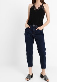 Svart spetstrimmad linne, löst sittande, i kombination med högmidjade mörkblå denimjeans. Skorna är svarta öppna mules med dekorativa detaljer.