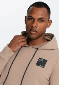Felpa beige con cappuccio, dotata di cordino, dettagli neri e logo quadrato sul petto. Tessuto morbido con una vestibilità comoda.