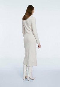 Robe à manches longues, côtelée, en beige clair, avec une silhouette ajustée et une longueur mi-mollet. Associée à des bottes blanches à talons blocs montant jusqu'aux genoux.