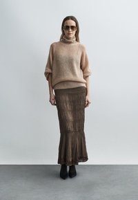 Ανοιχτό καφέ πλεκτό πουλόβερ με ψηλό γιακά, συνδυασμένο με μακρύ, πολύστιχο καφέ φόρεμα με τελείωμα με φούντες και μαύρα παπούτσια.