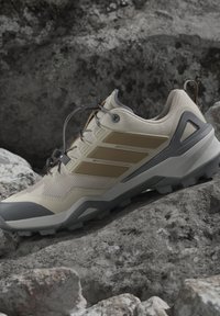 Scarpe da trail running beige e grigie con lacci sicuri, suola con battistrada e punta rinforzata, appoggiate su un terreno roccioso.