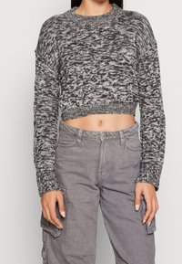 Avklippt svartvit stickad tröja med långa ärmar och vid halsringning, i kombination med grå cargobyxor med fickor på sidan.