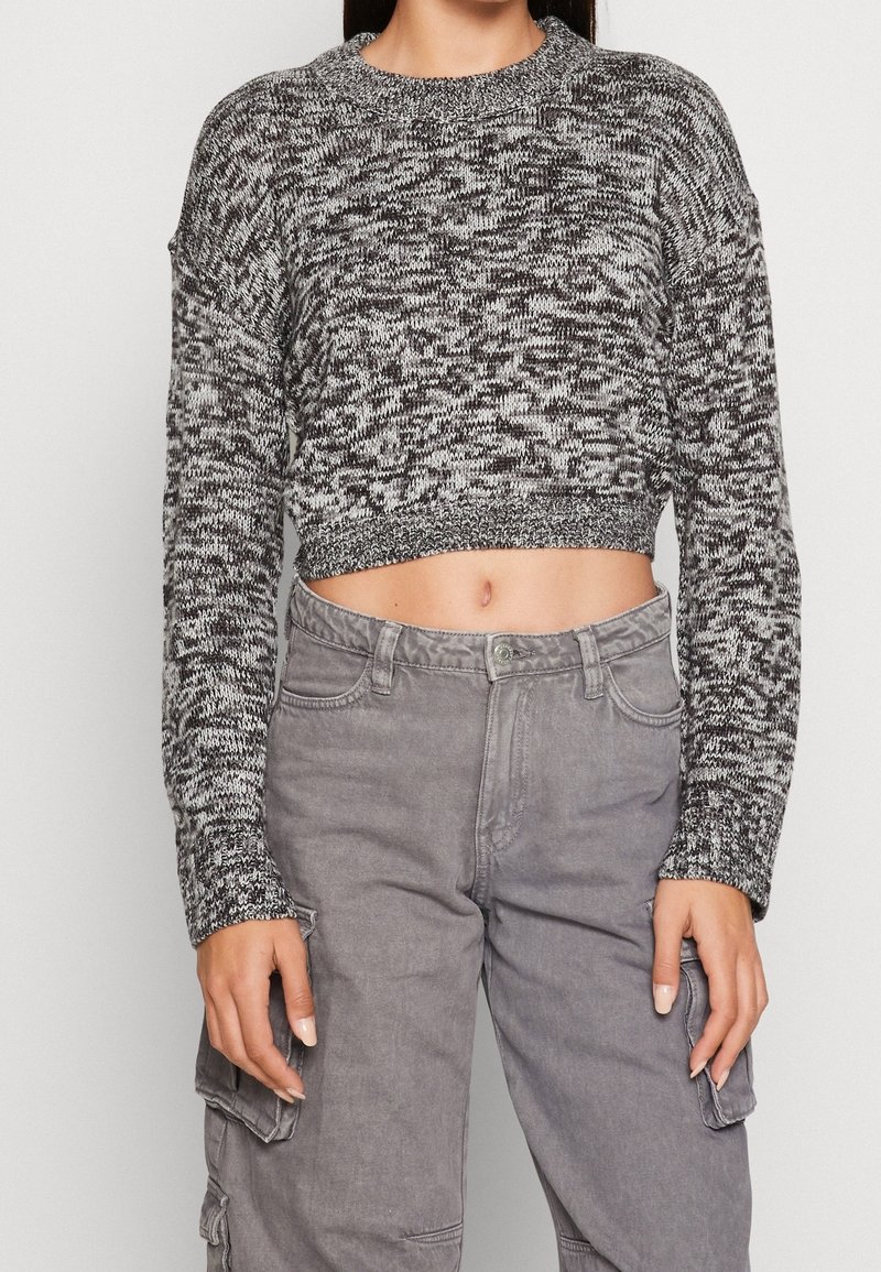 Avklippt svartvit stickad tröja med långa ärmar och vid halsringning, i kombination med grå cargobyxor med fickor på sidan.