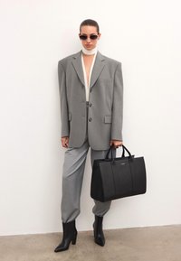 Szürke, oversized blézer, hozzáillő nadrággal, fehér felsővel és fekete, strukturált táskával. A modellen napszemüveg és hegyes, fekete magassarkú csizma van.