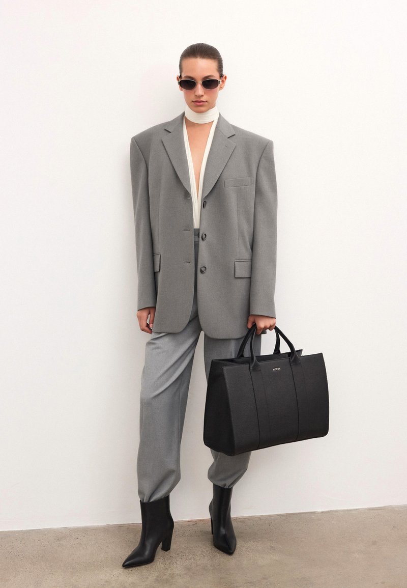Szürke, oversized blézer, hozzáillő nadrággal, fehér felsővel és fekete, strukturált táskával. A modellen napszemüveg és hegyes, fekete magassarkú csizma van.