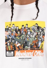 White cotton t-shirt featuring a colorful graphic print of a crowd scene, a footballer jumping, and "Weekend Offender" text.