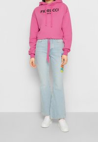 Sudadera rosa recortada con logo negro "Fiorucci", combinada con vaqueros acampanados azul claro con parches coloridos. Zapatillas blancas completan el conjunto.