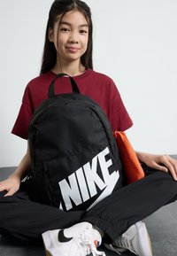 Juodas Nike kuprinės su dideliu baltu logotipu, pagaminta iš lygios audinio tekstūros ir užtrauktukų; šalia matomas oranžinis maišelis.