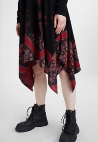 Robe noire avec des motifs floraux rouges et géométriques, ourlet asymétrique, associée à des bottes noires à lacets avec une finition texturée.