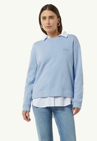 Lichtblauwe sweatshirt met geborduurde tekst, gelaagd over een wit overhemd met kraag. Kenmerkt een relaxte pasvorm en ribgebreide manchetten. Spijkerbroek zichtbaar.