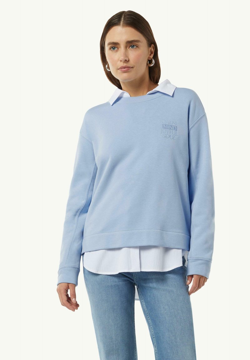 Lichtblauwe sweatshirt met geborduurde tekst, gelaagd over een wit overhemd met kraag. Kenmerkt een relaxte pasvorm en ribgebreide manchetten. Spijkerbroek zichtbaar.