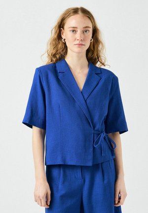 Blazer envolvente de lino azul con mangas cortas, que presenta un cierre de lazo y un cuello con muescas. Textura suave y diseño ajustado.