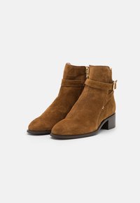 Hnědé semišové ankle boots s mírně špičatou špičkou, krátkým blokovým podpatkem a zlatými kovovými detaily. Obsahují zip a pásek.