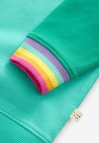 Felpa turchese con polsini a coste multicolore che presentano strisce viola, rosa, gialle e verdi. L'etichetta dice "Little Bird".