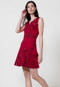 Rotes ärmelloses Kleid mit schwarzer floraler Stickerei, tailliertem Oberteil und schwingendem Rock. Glatte Stoffoberfläche mit leichtem Glanz und filigranem Muster.