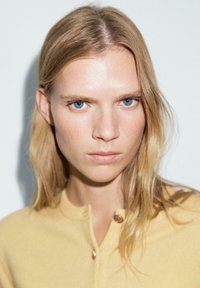 Porträtt av en person med långt, vågigt blont hår, iförande en gul knapp-up tröja med mjuk textur och rundad halsringning.