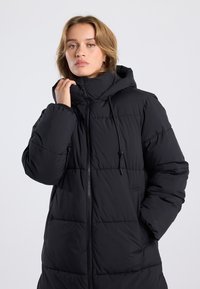 Black padded coat with a high collar and adjustable hood. Features a front zipper, elastic cuffs, and side pockets. Textured fabric, quilted design.