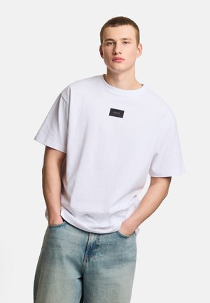 Młody mężczyzna z krótkimi włosami, ubrany w zwykłą białą, oversize'ową koszulkę z małą czarną naszywką z logo i jasnoniebieskie wyblakłe jeansy, ręce w kieszeniach.