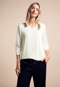 Blusa bianca a V con maniche a tre quarti, texture liscia, abbinata a pantaloni scuri, su uno sfondo neutro.