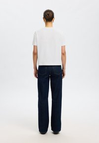 Femme avec les cheveux en chignon portant un t-shirt blanc uni à manches courtes et un jean bleu foncé droit, vue de dos sur un fond uni.