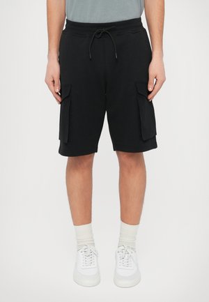 Shorts cargo en coton noir avec une taille à cordon, des poches latérales et une coupe décontractée, assortis à des baskets blanches et des chaussettes claires.