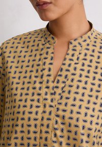 Geel shirt met een mandarin kraag, voorzien van een paisleypatroon in marineblauw en wit. Zachte textuur met een knoopsluiting.
