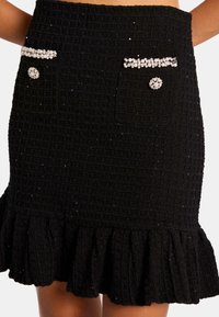 Jupe noire texturée avec un ourlet volanté, deux poches avant ornées de strass et un motif en quadrillage sur toute la surface du tissu.