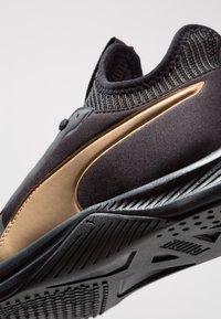 Zapatilla deportiva negra con un diseño elegante y un detalle dorado, superior de punto y suela texturizada para agarre. Material suave con detalles añadidos.