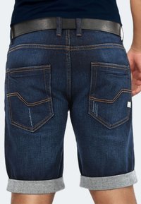 Jeansshorts mit einem dunkelblauen Waschung, umgeschlagenen grauen Säumen und zwei Gesäßtaschen, die mit orangefarbenen Nähten und einem kleinen Etikett hervorgehoben sind.