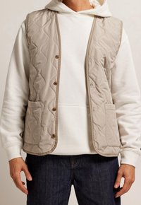 Beżowa pikowana kamizelka z zaokrąglonym dołem, zapinana na pięć guzików, z dwoma przednimi kieszeniami. Nosi się ją na białą bluzę z kapturem i ciemne dżinsy.