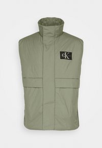 Prošívaná vesta ve světle olivové barvě s vysokým límcem, dvěma předními kapsami a černou logovou nášivkou na hrudi. Hladká, texturovaná látka.