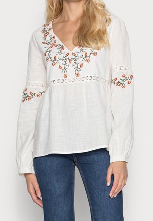 Nainen, joka pitää päällään valkoista pitkähihainen pusero, jossa on oransseja kukkaompeleita rinnassa ja hihoissa, yhdistettynä sinisiin farkkuihin.