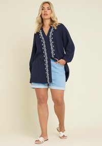 Blusa blu navy con bottoni, decorata con ricami geometrici bianchi sulla parte anteriore, maniche lunghe e vestibilità rilassata, abbinata a pantaloni corti di jeans chiari.