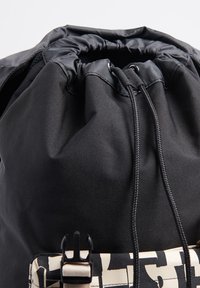Mochila negra con cordón, que presenta una parte superior fruncida, un tejido duradero y un acento de patrón beige en contraste en la parte delantera.