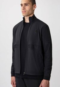 Black jacket made of lightweight fabric, featuring mesh panels at shoulders, zip front, high collar, and a hexagonal logo on the chest.