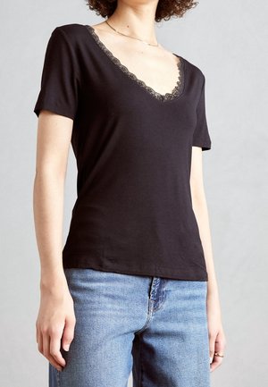 Vrouw draagt een zwart T-shirt met korte mouwen en V-hals met kanten rand en lichtblauwe spijkerbroek tegen een effen grijze achtergrond.