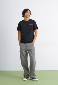 Sort t-shirt med korte ærmer, der har et lille logo på venstre bryst, parret med løse grå cargo-bukser og sort-hvide sneakers.