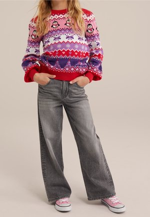 Pige iført en festlig sweater med pingviner og vintermønstre, grå vidde bukser og lyserøde sneakers, stående med hænderne i lommerne.